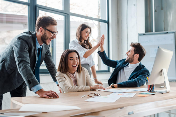 Four coworkers smile and high-five around a desk in a bright office.