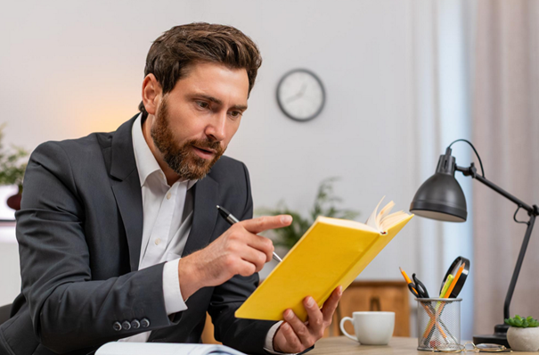 Man in a suit pointing with a pen while reading a yellow notebook at a desk