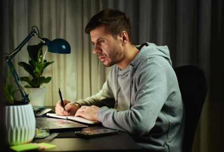Man in a gray hoodie writing in a notebook at a desk