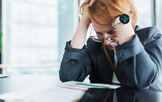 A tired professional holds her head at a desk, illustrating the strain of excessive pressure
