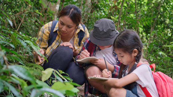 An adult and two children observe plants and take notes in a forest.